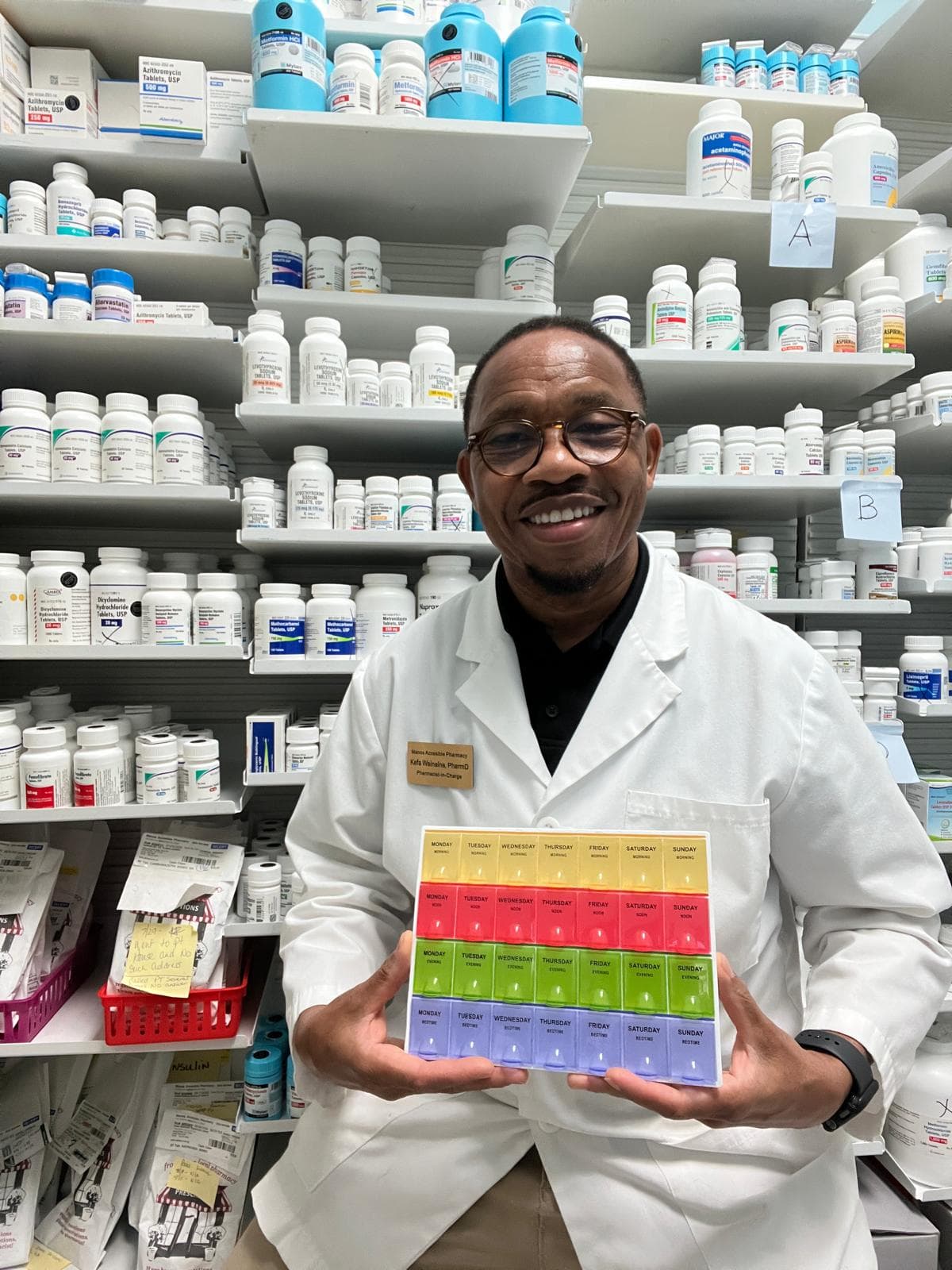 A pharmacist in a white coat stands before the dispensing shelves holding out a seven-day organizer, its four rows running yellow Morning, red Noon, green Evening, and purple Bedtime.