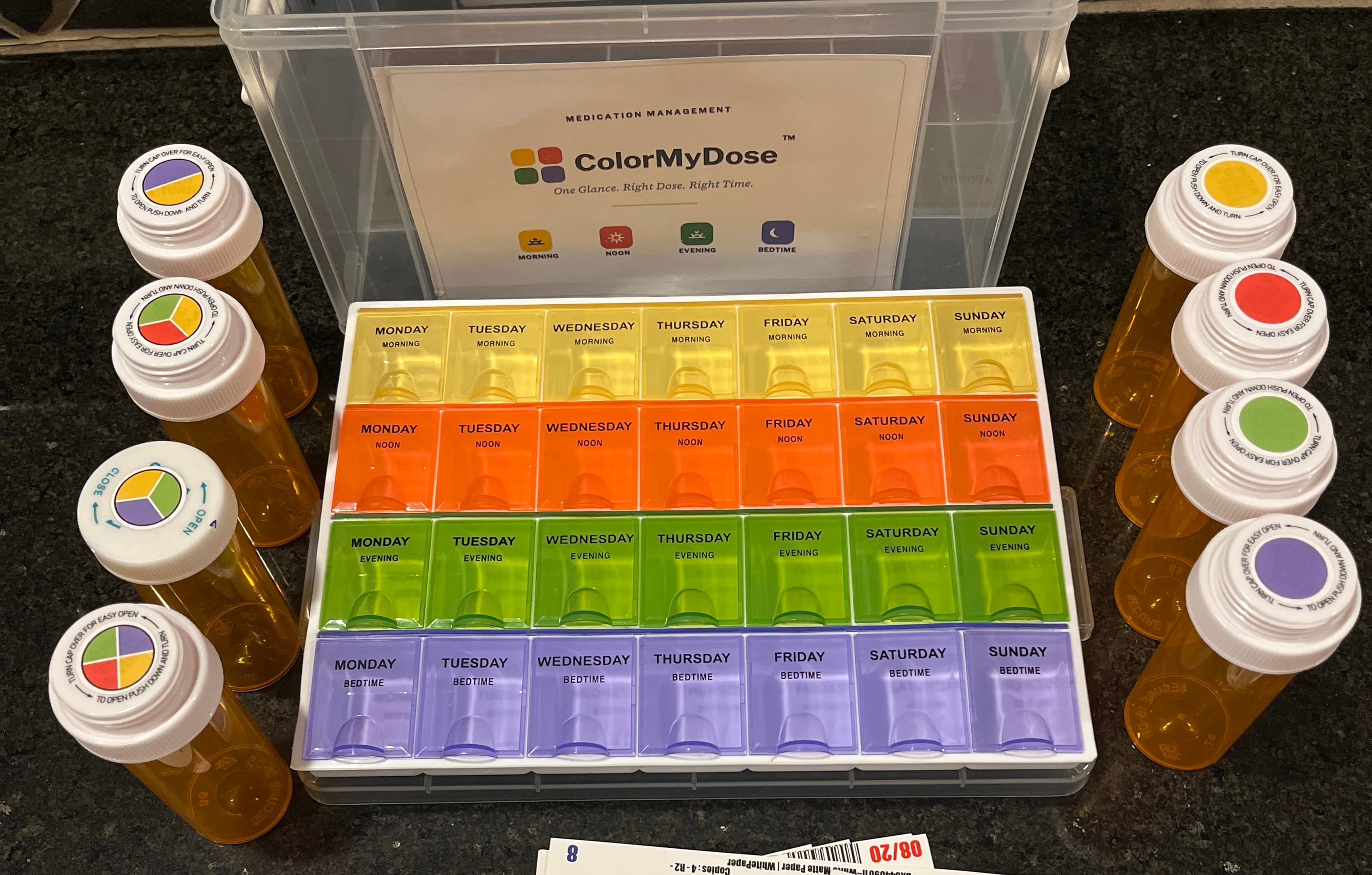 A ColorMyDose kit on a counter: a seven-day organizer with yellow Morning, orange Noon, green Evening, and purple Bedtime rows labeled Monday through Sunday, flanked by prescription bottles wearing matching color-coded caps.