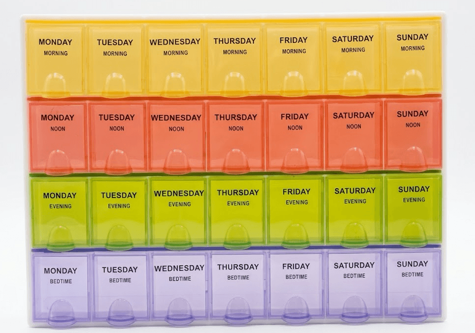 A seven-day organizer photographed from above, its four rows running yellow Morning, red Noon, green Evening, and purple Bedtime, each compartment labeled Monday through Sunday.