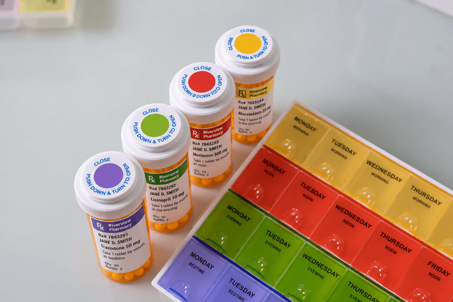A ColorMyDose kit on a counter: a seven-day organizer with yellow Morning, red Noon, green Evening, and purple Bedtime rows labeled Monday through Sunday, beside prescription bottles whose caps and label bands carry the matching colors.