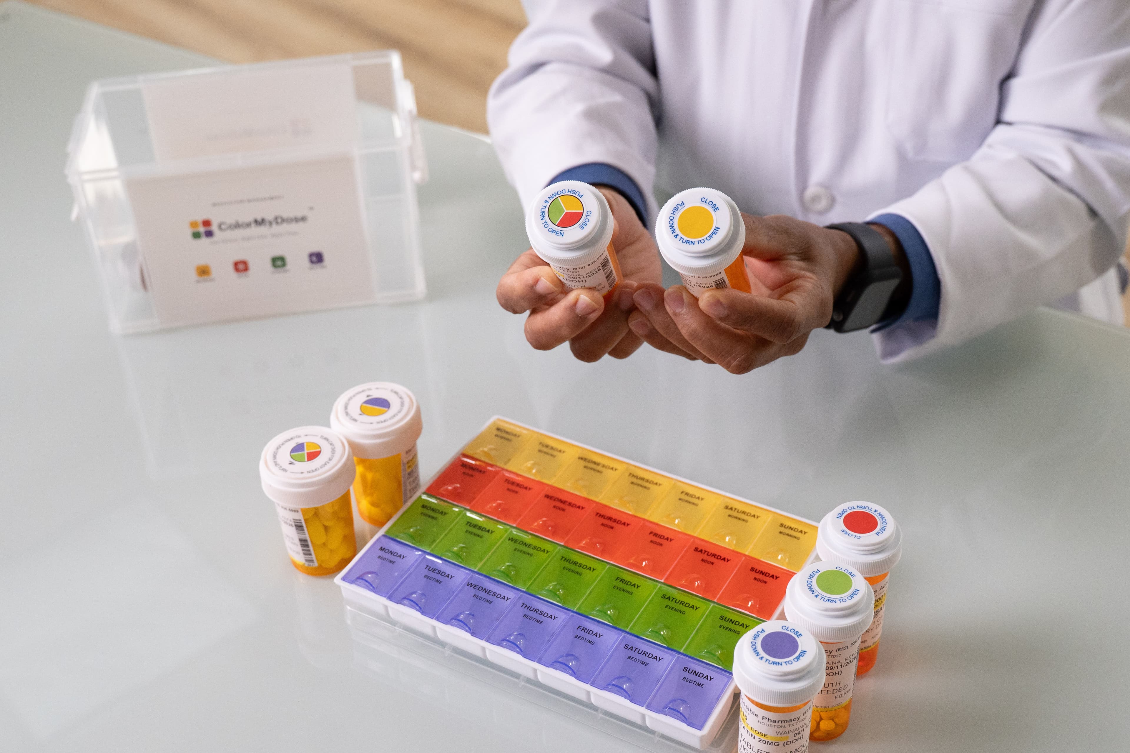 Prescription bottles on a counter surrounded by their caps: single-color caps in yellow Morning, red Noon, green Evening, and purple Bedtime, alongside split caps that pair two colors for medications taken more than once a day.
